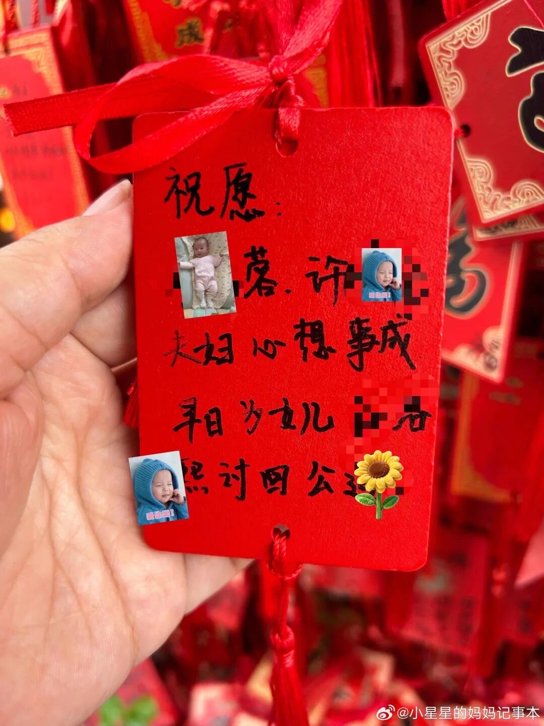 手持一块红色祈福牌，手写黑字祝愿这对夫妇心想事成、为女儿讨回公道，人名处被贴纸和马赛克遮盖，牌上另贴着三张婴儿照片